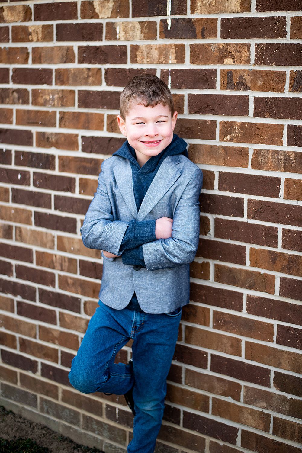 A smiling child in a gray blazer and blue jeans poses casually against a brick wall background.
