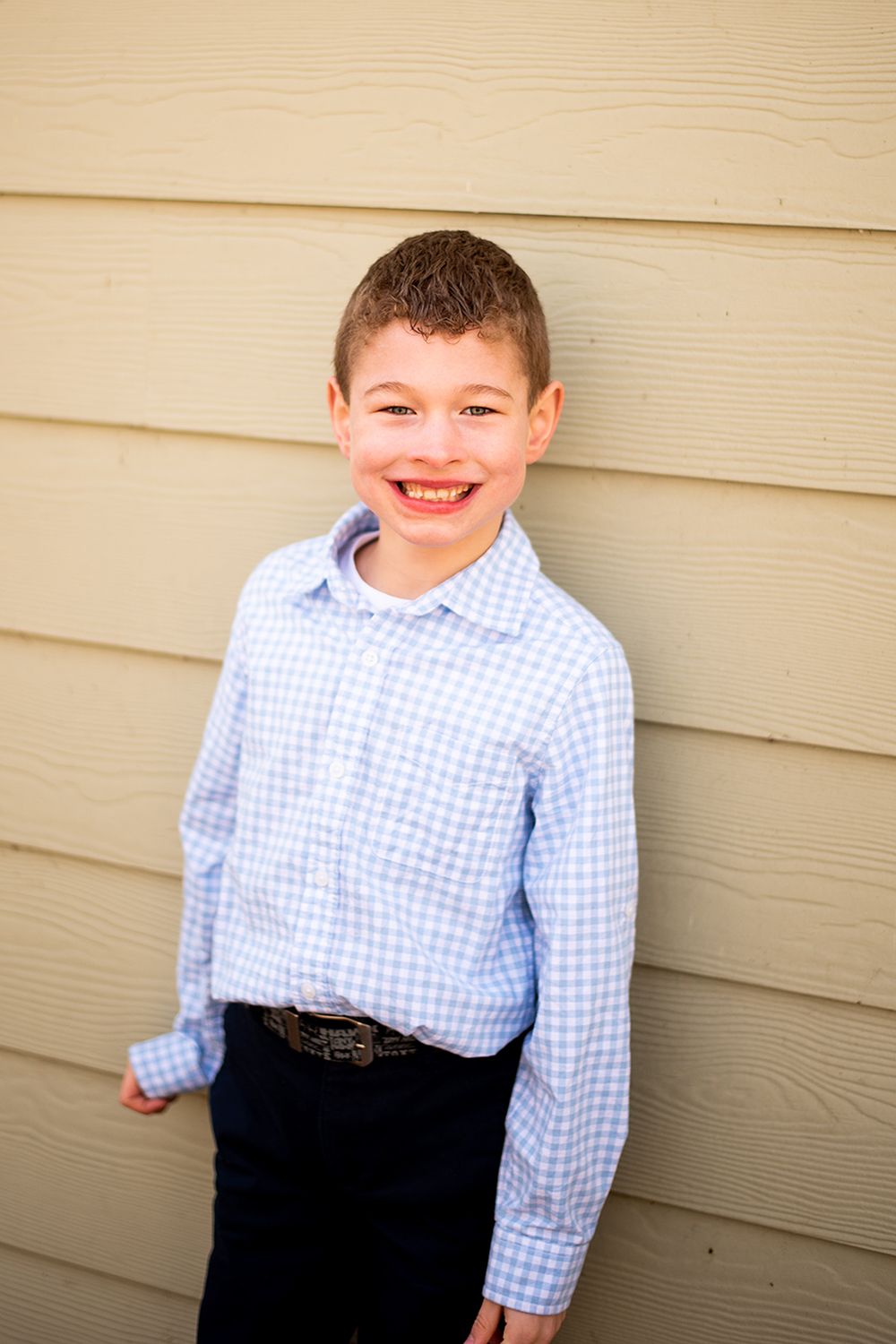 Young boy in blue gingham shirt and dark pants posed against beige siding outdoors.