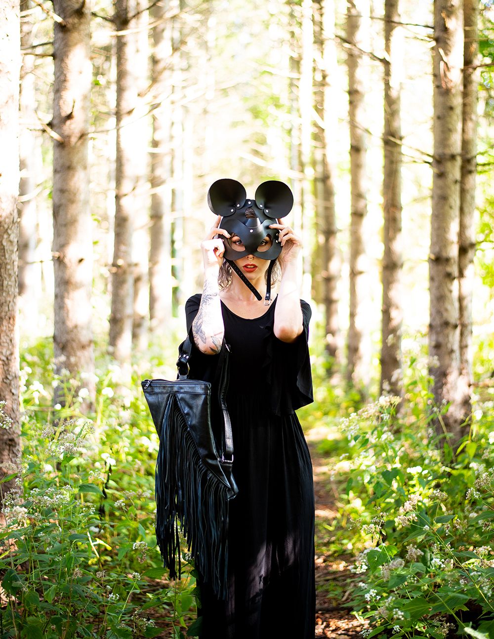 A person in a black dress and mouse ears stands in a sunlit forest holding a camera and fringe bag.
