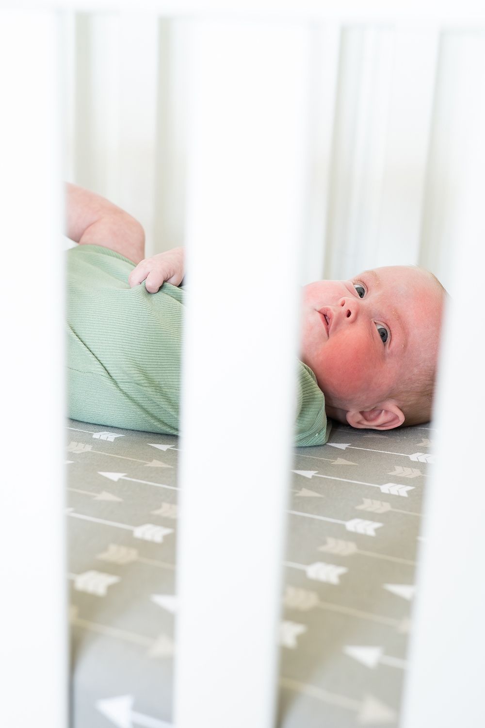 Baby lying on a polka dot crib sheet wearing mint green sleepwear, viewed through white crib bars.