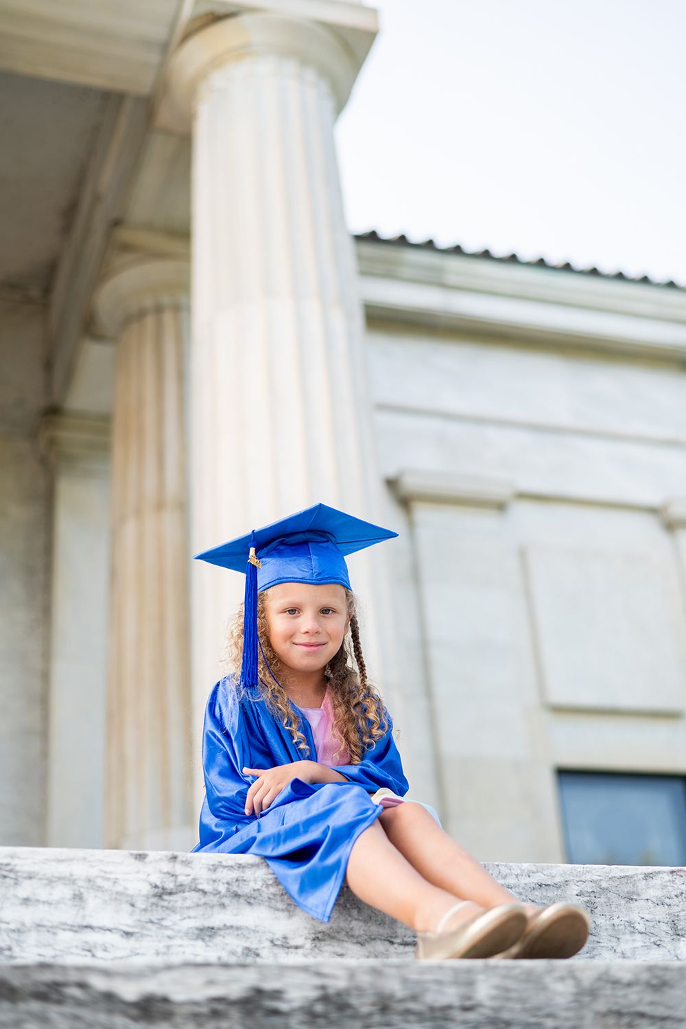 Graduate in blue regalia sits on stone ledge in front of classic white pillars during graduation photoshoot.