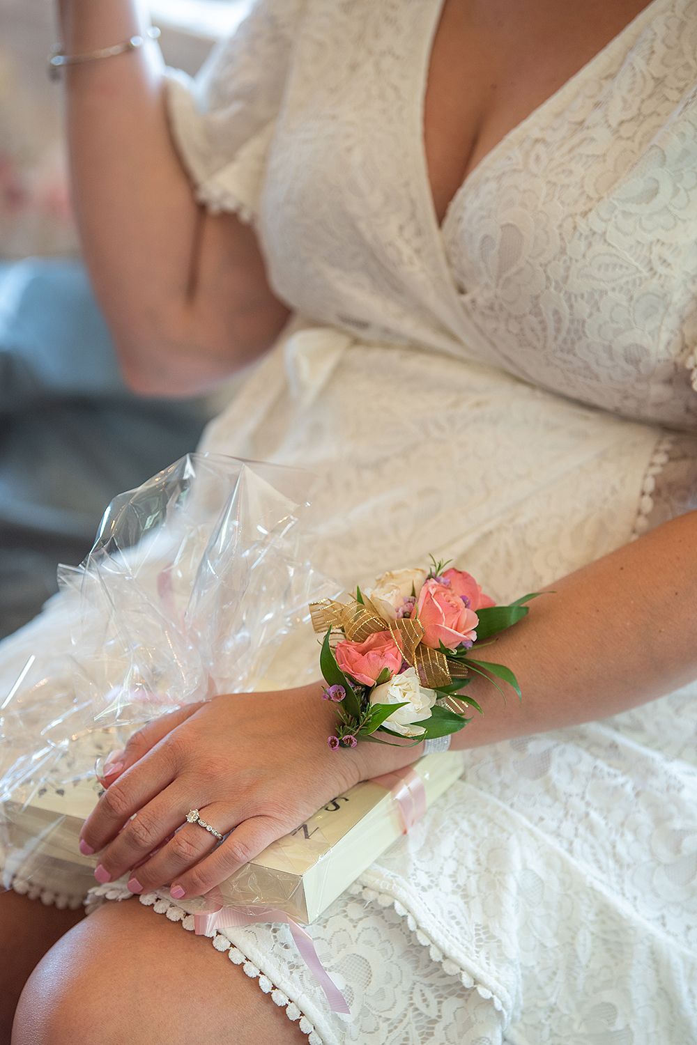 Close-up shot of hands wearing a delicate lace wedding dress with pink floral corsage and engagement ring.