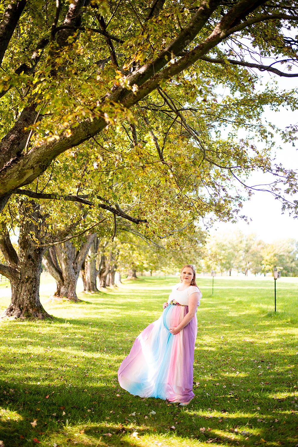 A person in a flowing pastel dress twirls gracefully beneath a row of autumn trees in a sunlit park.