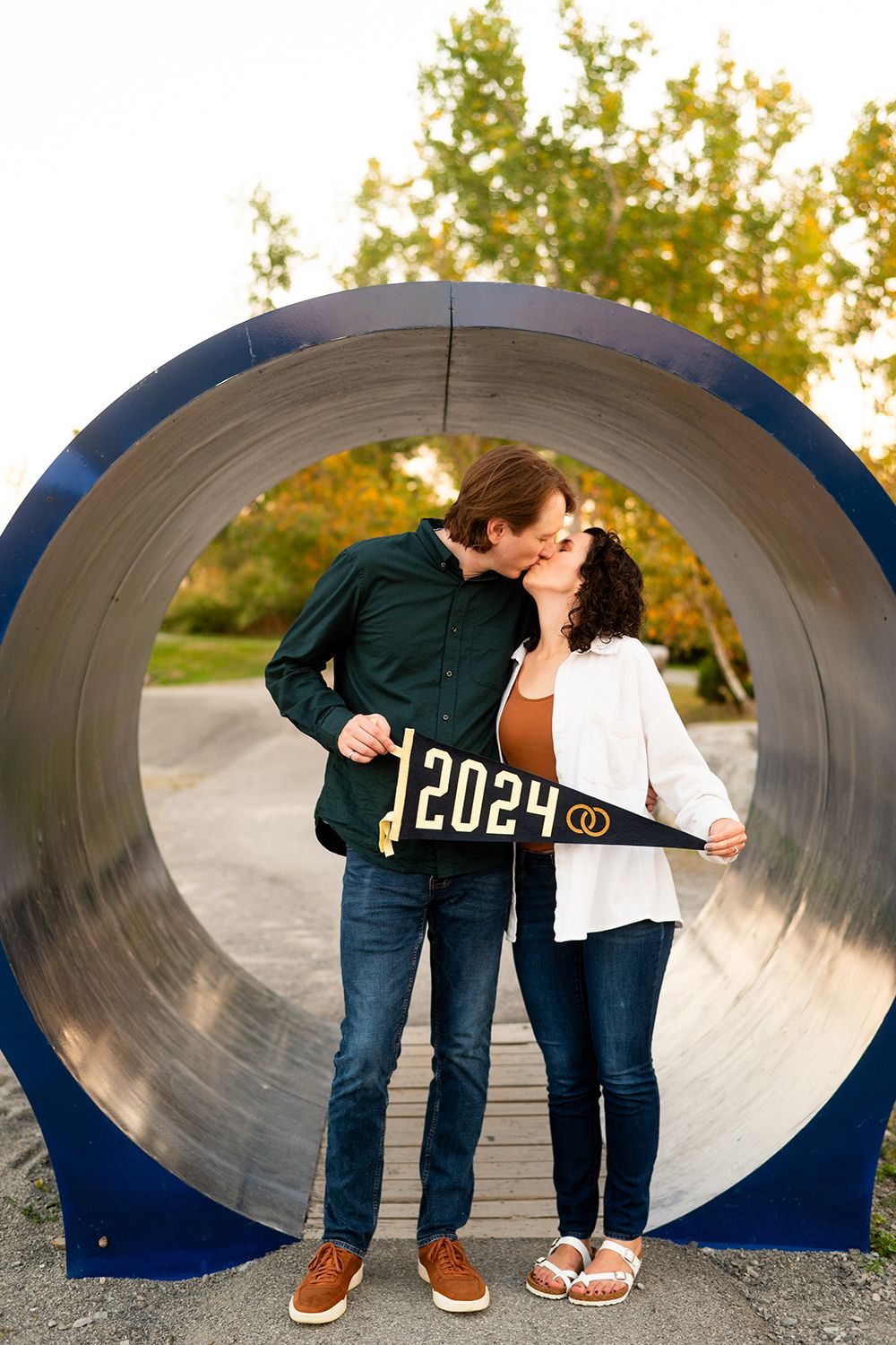 A couple shares a romantic moment while holding a 2029 sign inside a large circular tunnel during an autumn day.