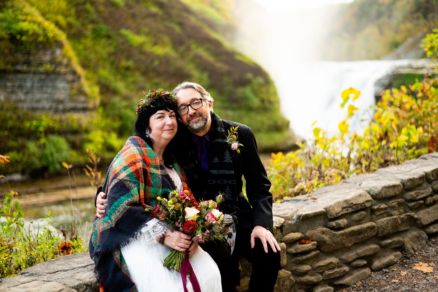 A couple sits together on stone wall with autumn foliage and mountains in background holding vibrant floral bouquet.