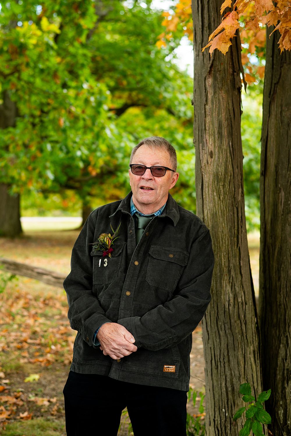 Someone wearing sunglasses and a dark jacket stands smiling near autumn trees in a park.
