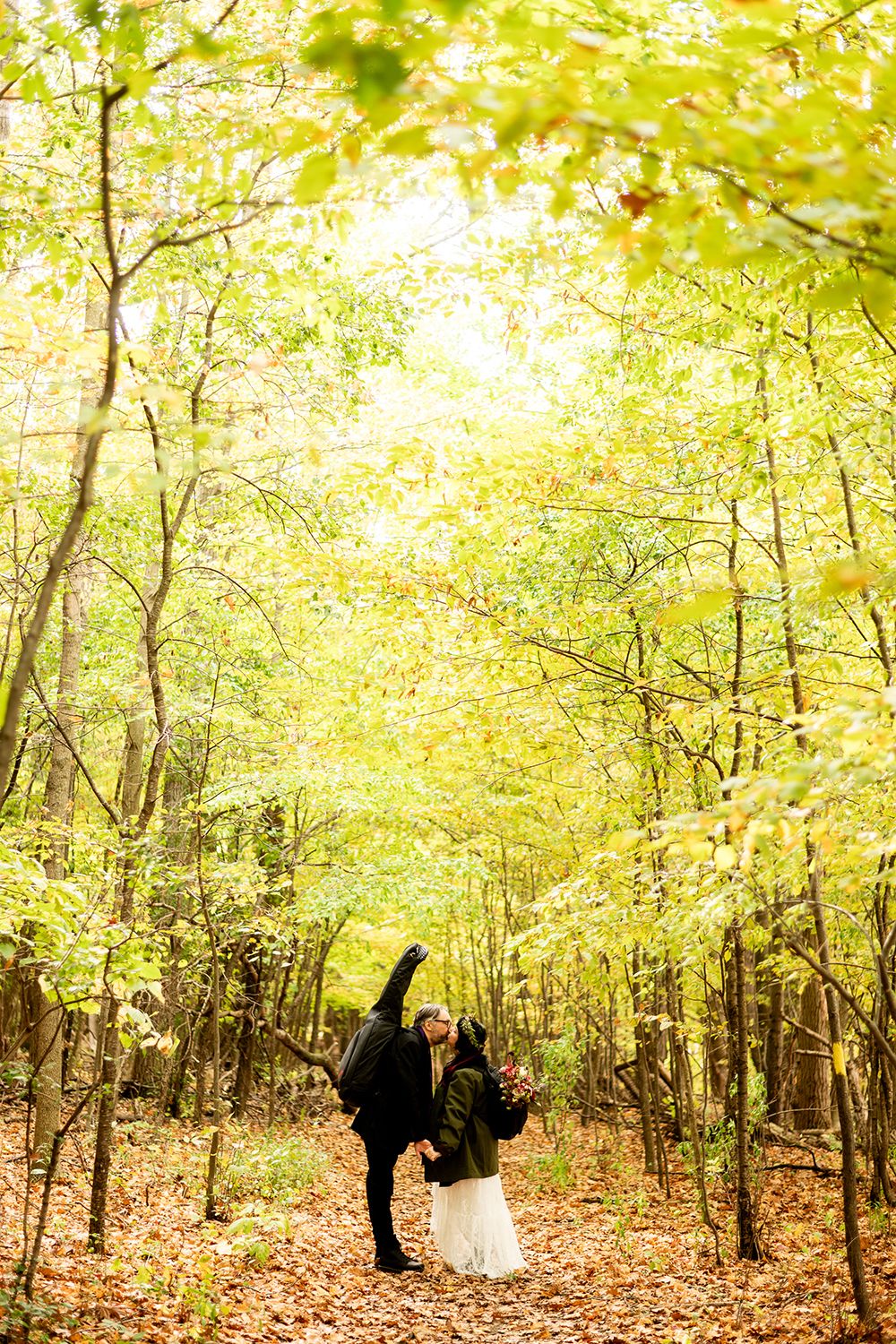 Couple walking together through a sunlit autumn forest with golden yellow foliage overhead on a path covered in fallen leaves.
