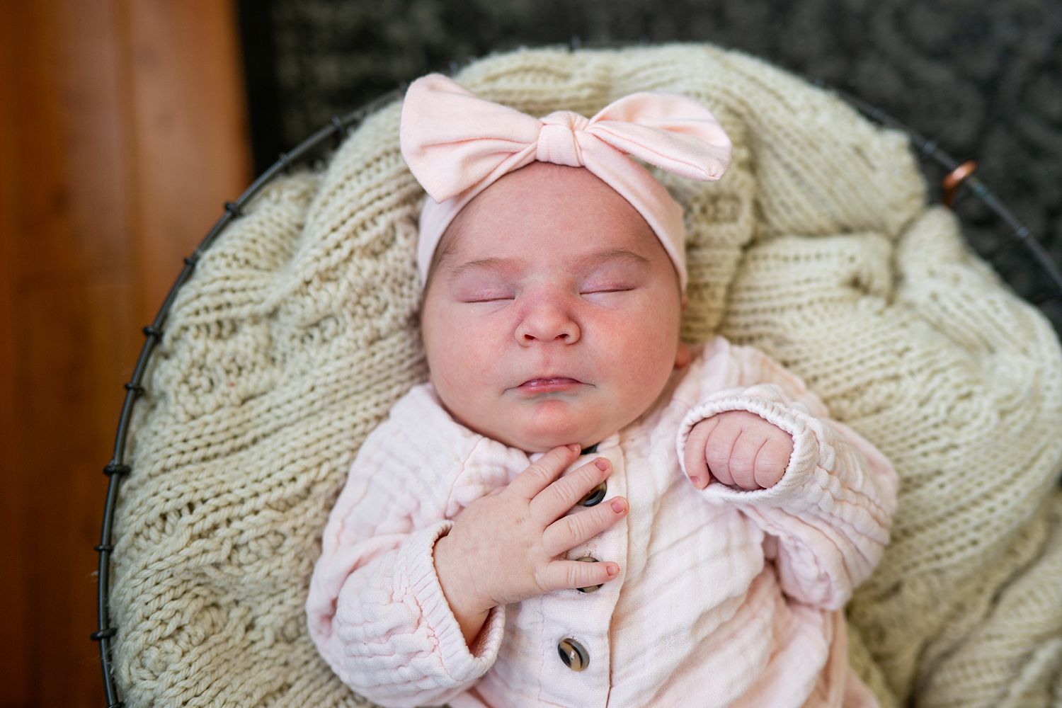 Newborn baby wearing pink bow sleeps peacefully wrapped in cozy knitted blanket.