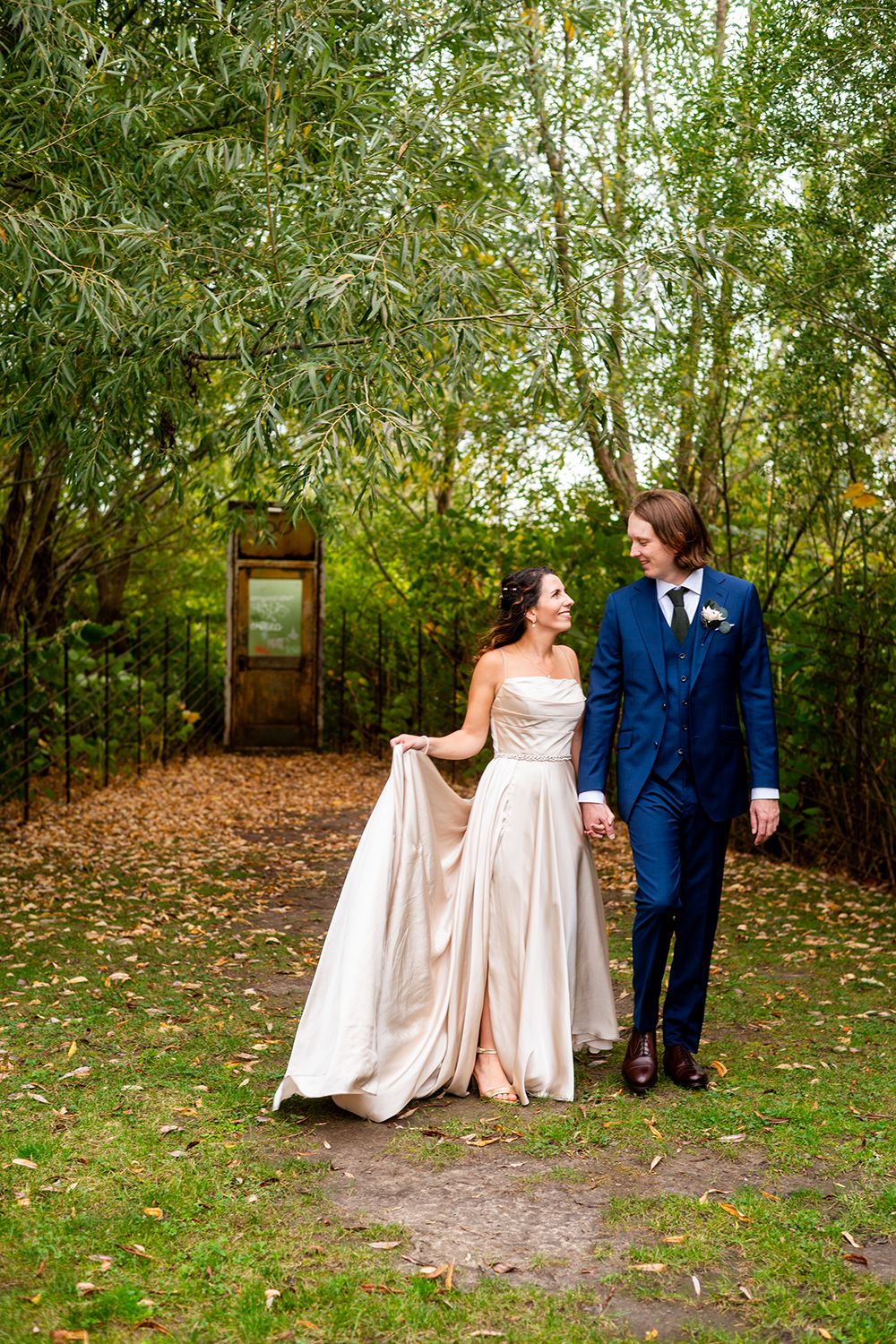 Couple in formal wedding attire strolls through wooded garden path with autumn leaves on ground.