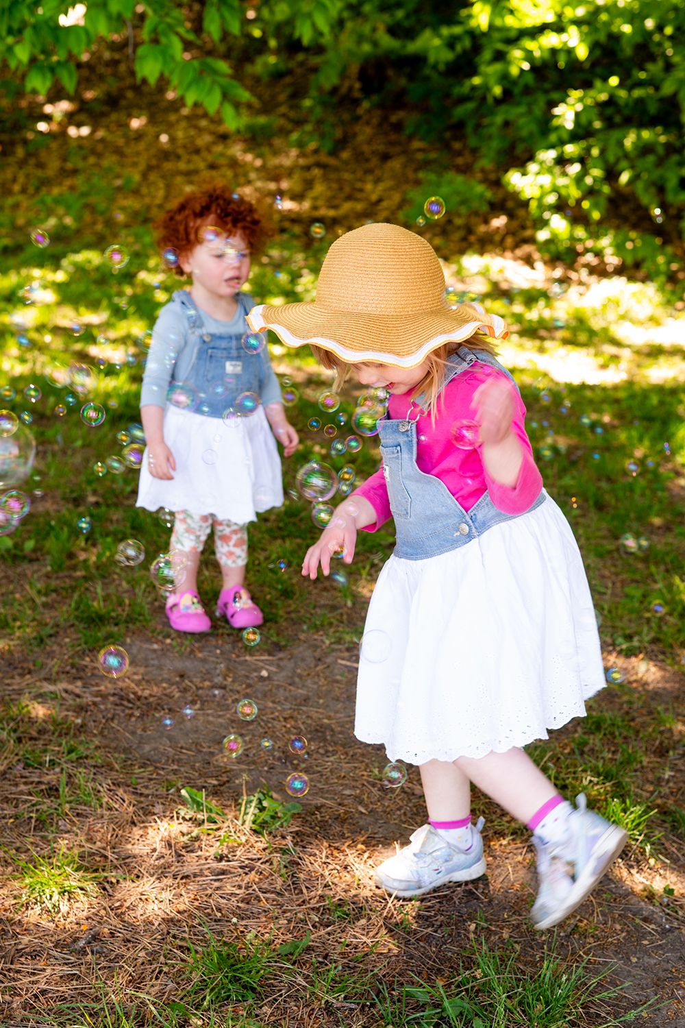 Two young children in denim outfits and white skirts playing with bubbles in a sunny wooded area on a summer day.