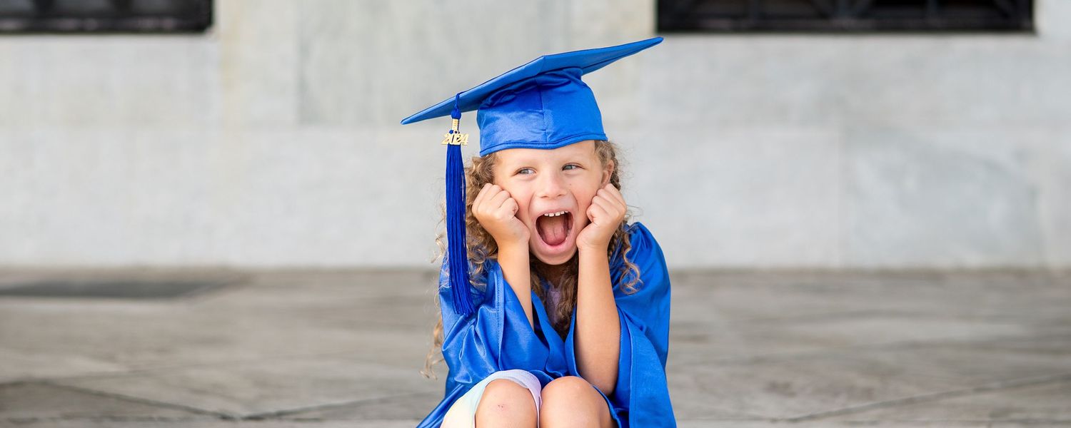 Graduate in blue cap and gown jumping with excitement during outdoor graduation celebration.