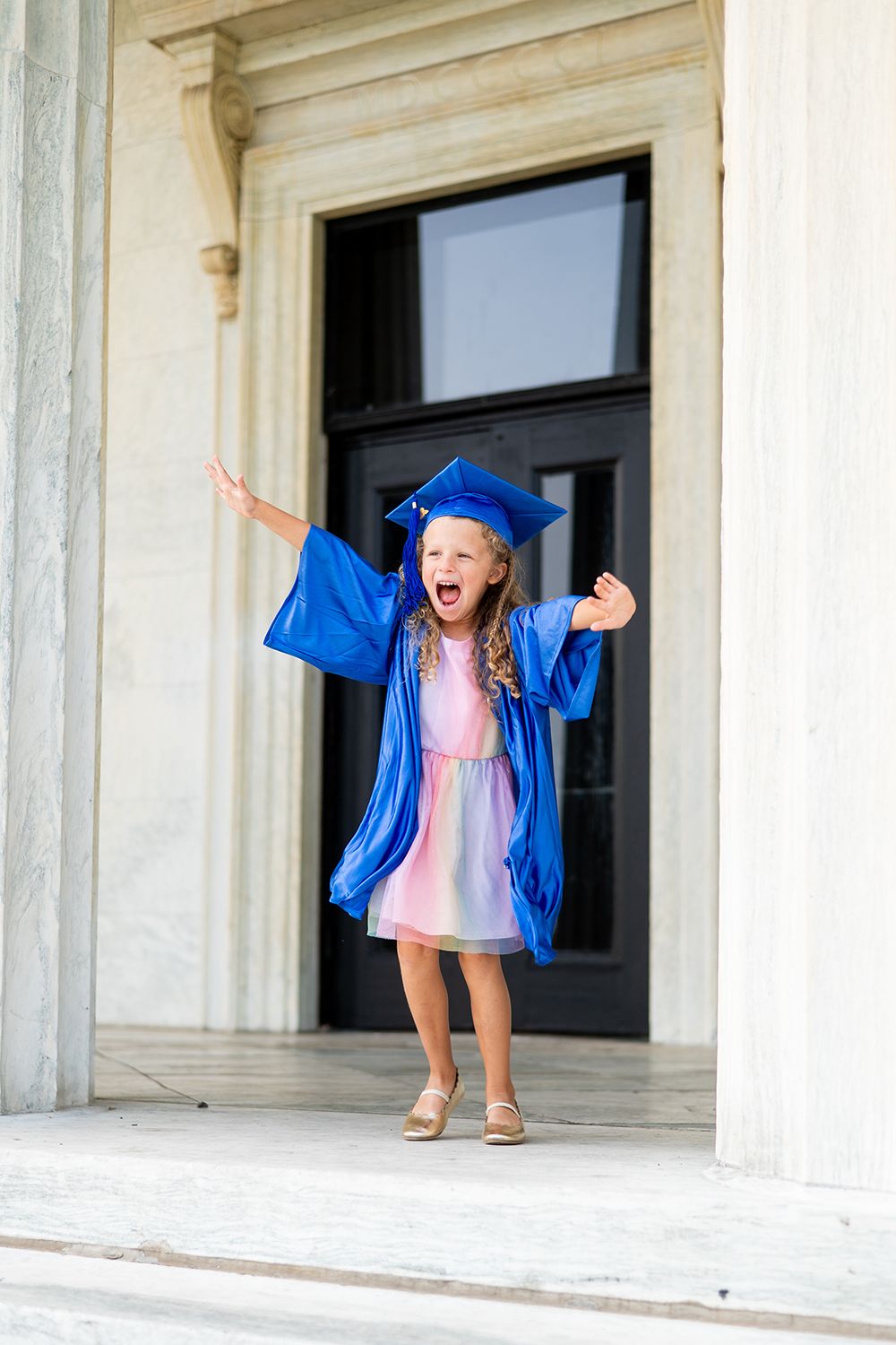 A jubilant graduate in bright blue cap and gown stands excitedly in a doorway of a classical building.