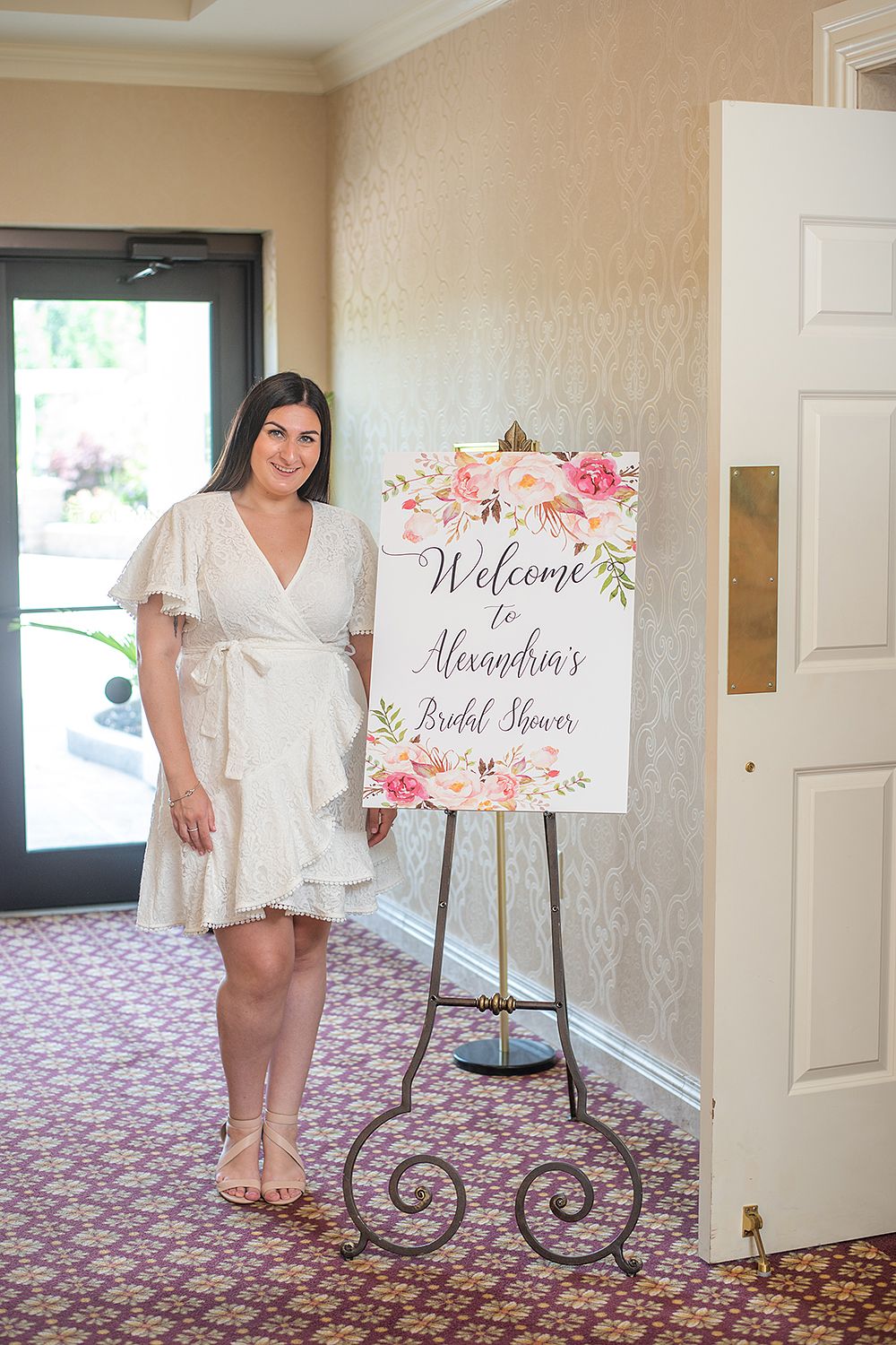 Floral welcome sign on an ornate easel stand next to an entryway with patterned carpet and beige walls.