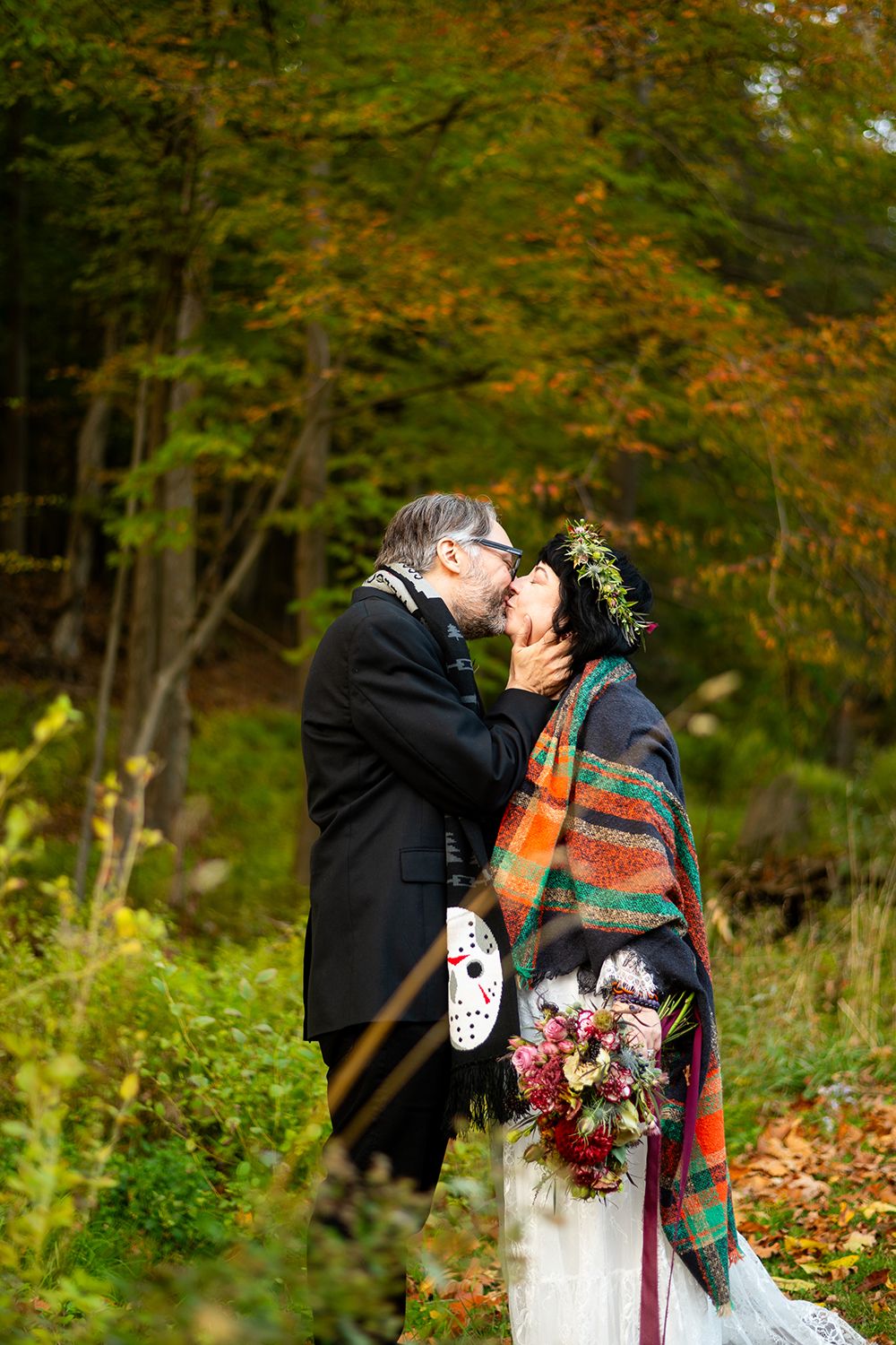 A couple shares a romantic kiss in an autumn forest setting with colorful fall foliage and a bridal bouquet.