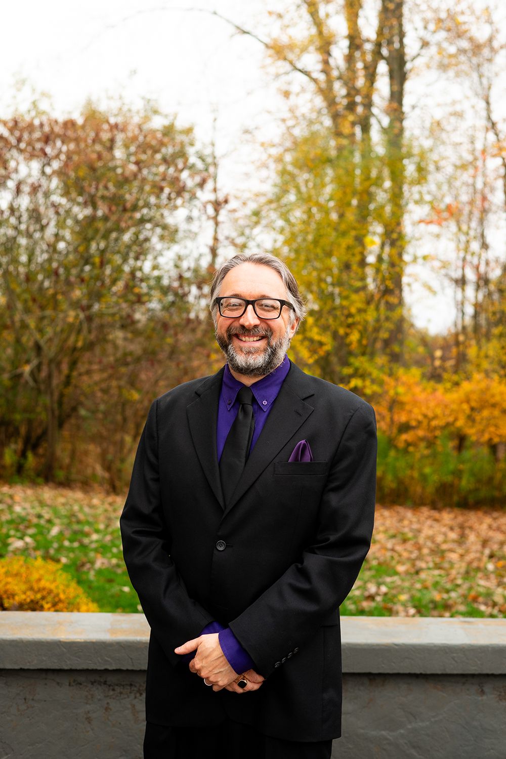 A person in a black suit and purple tie poses outdoors against an autumn backdrop with colorful fall foliage.