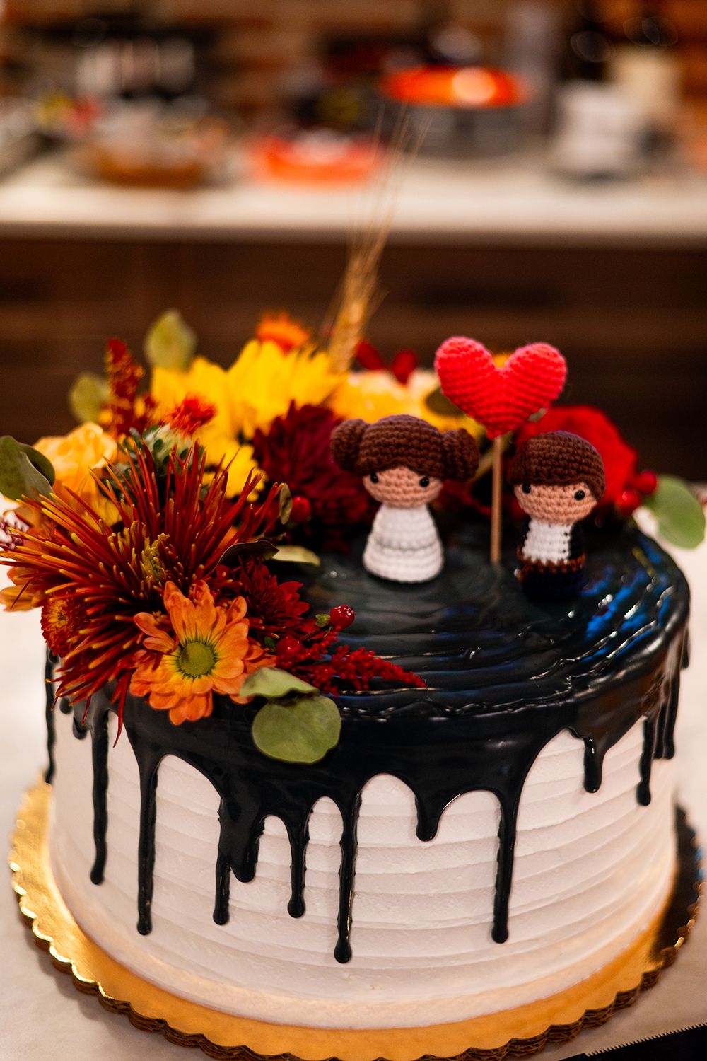 Wedding cake decorated with black drip frosting, fresh autumn flowers and small decorative figurines on top.