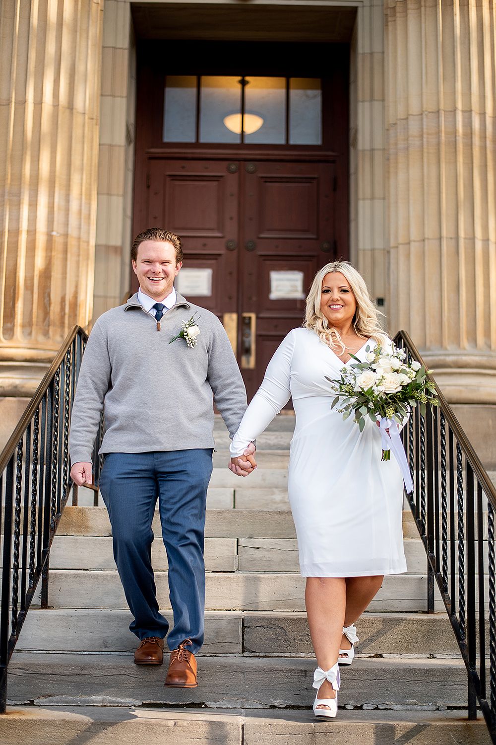 Modern wedding party walks down formal stone steps in coordinated gray and white attire holding hands.