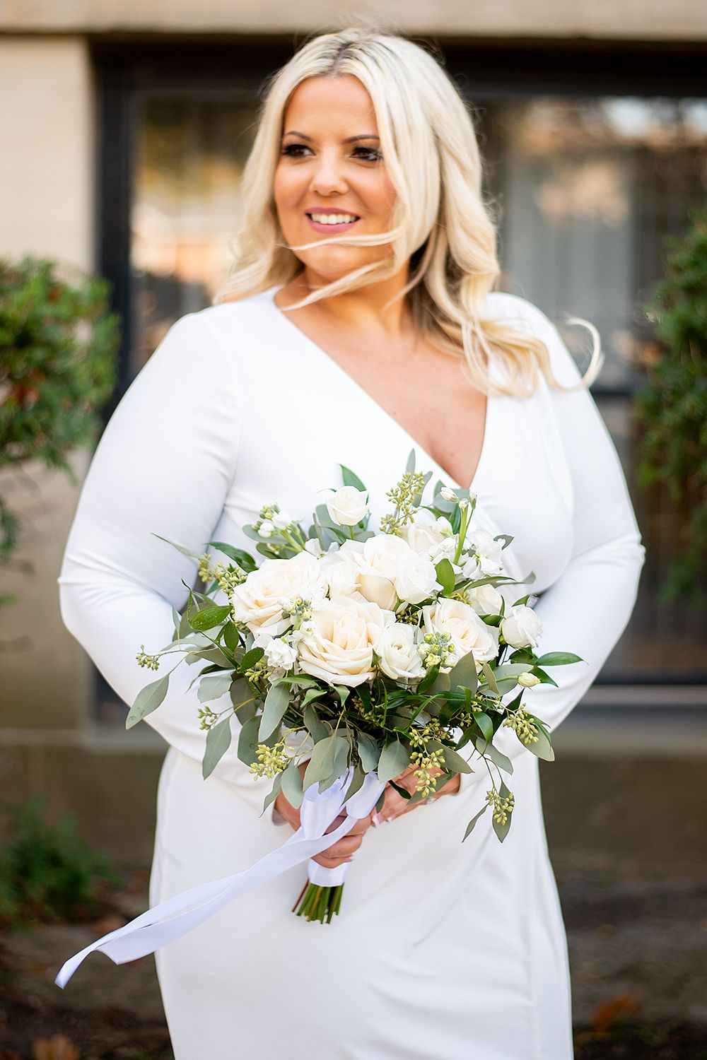 A bride in a white long-sleeved dress holds an elegant bouquet of white roses and greenery while posing outdoors.