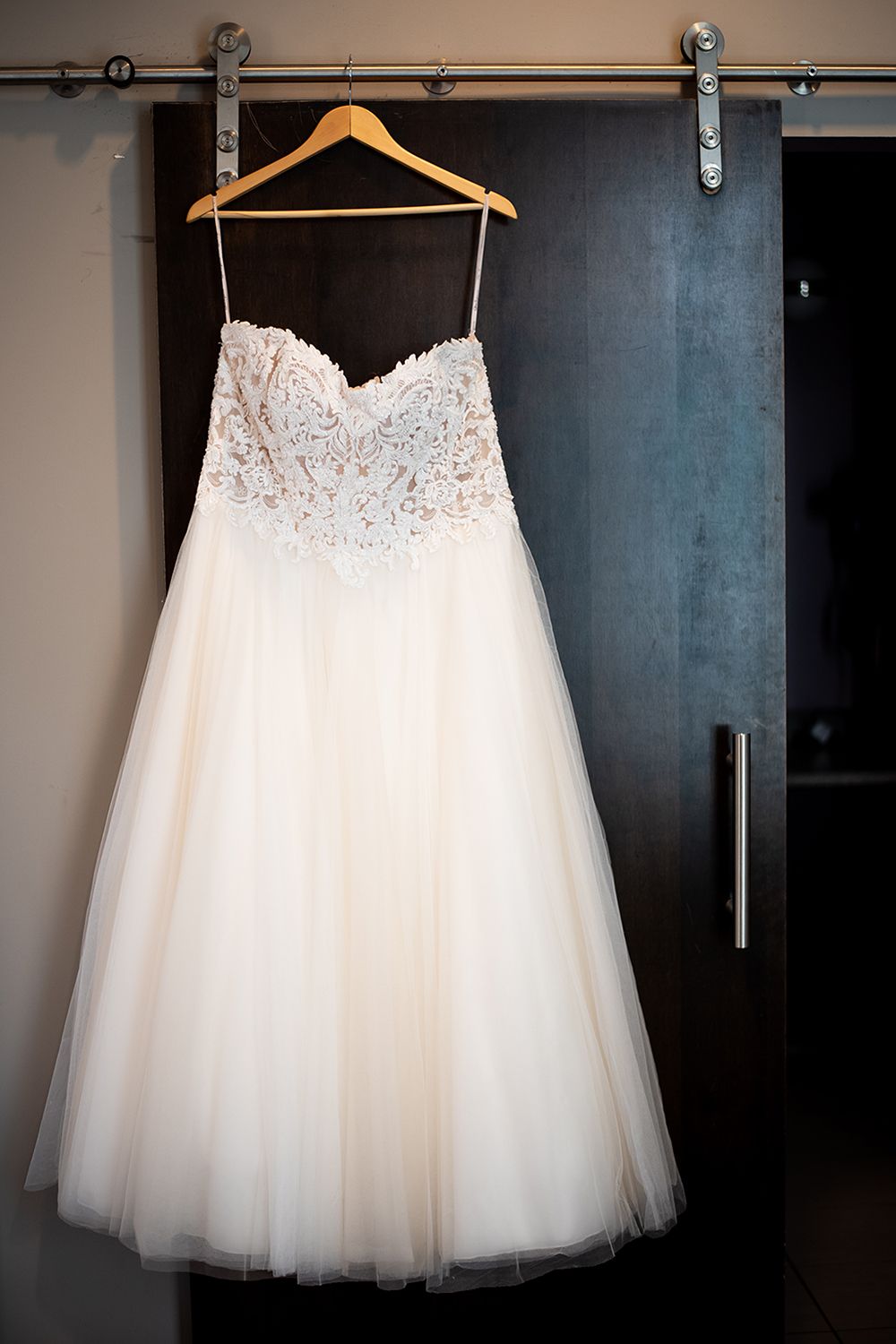 White strapless ball gown wedding dress with lace bodice and tulle skirt hanging on wooden hanger against dark wall.