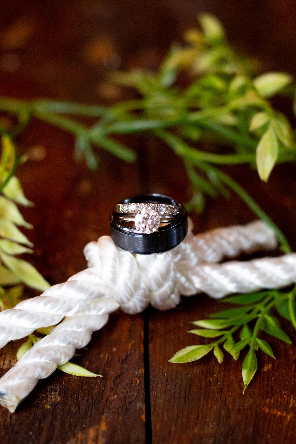 Diamond engagement ring with white ribbon and green foliage on rustic wooden surface with dramatic lighting.