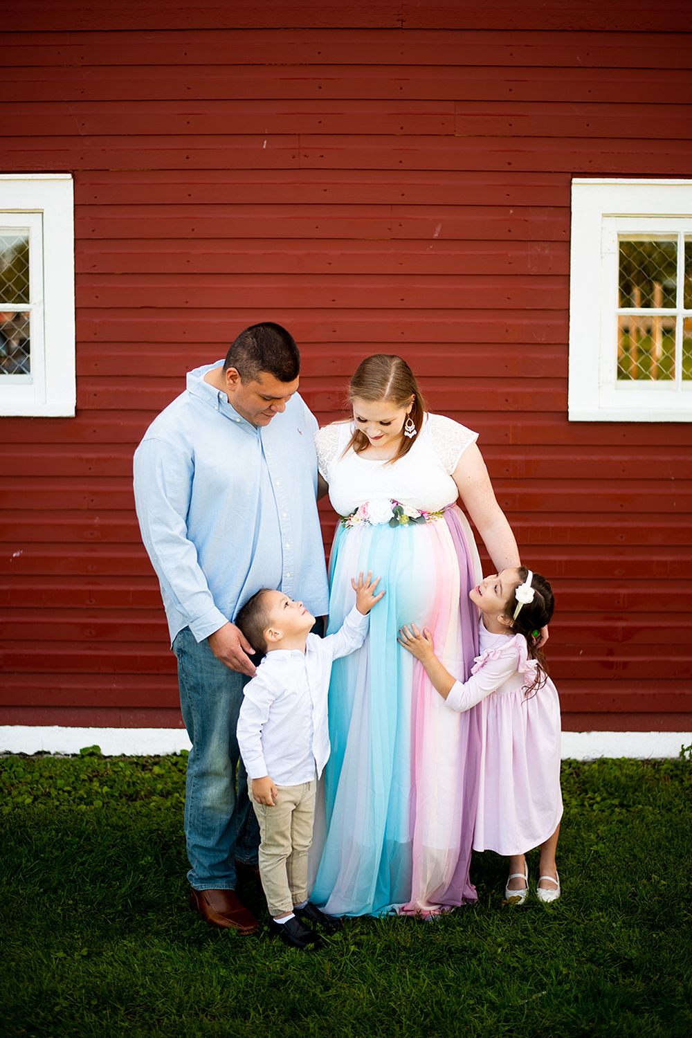 Sweet family moment captured against a red barn wall as they share affectionate poses on the grass.