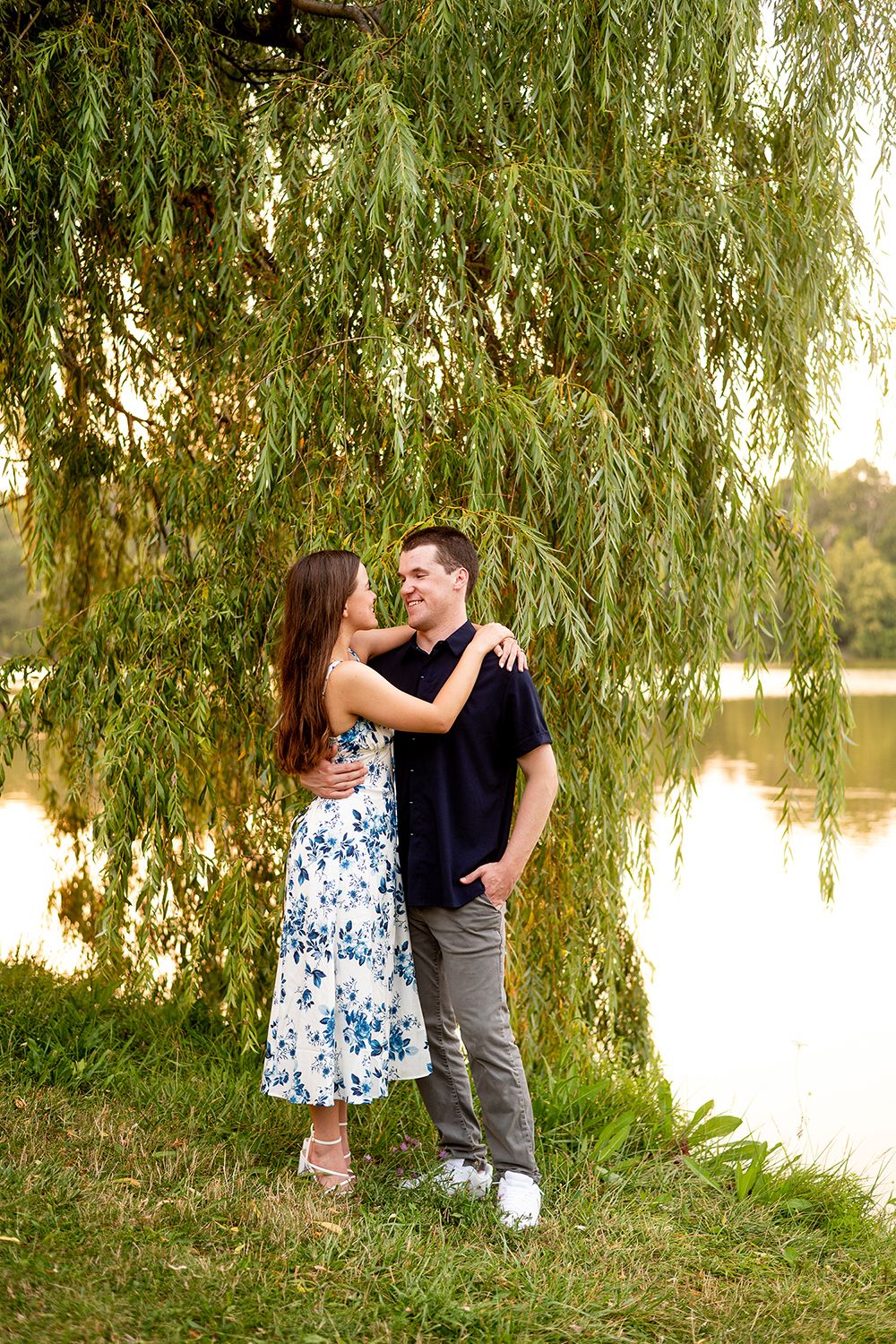 A couple embraces by a lake under a weeping willow tree during golden hour.
