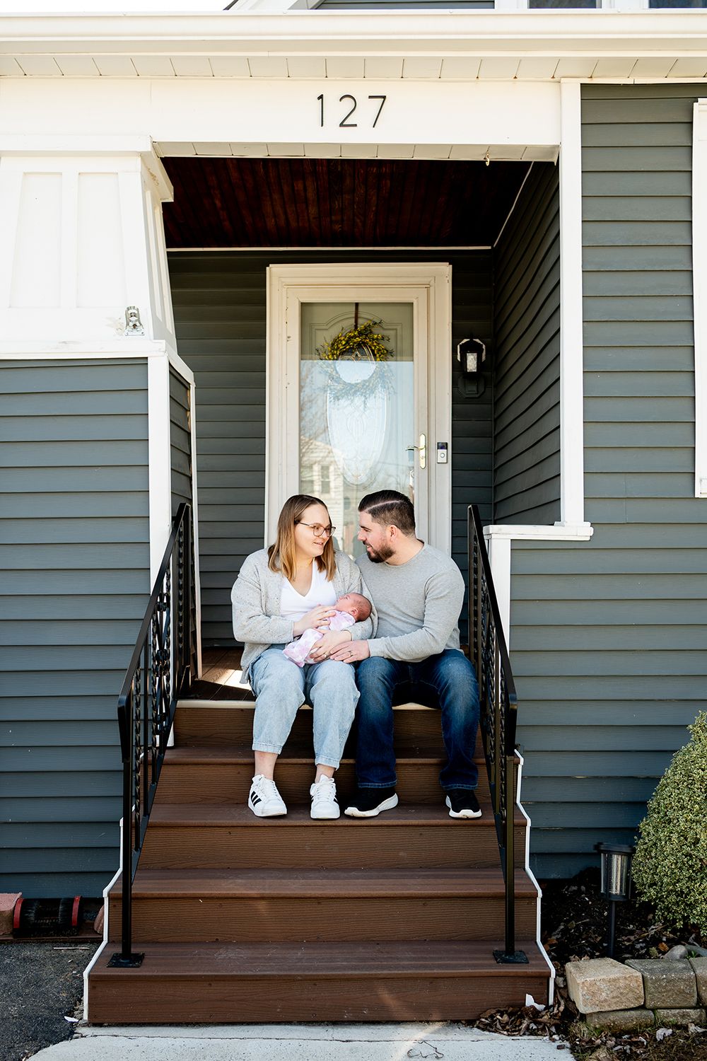A family sits together on the wooden steps of their gray house with the number 127 visible.