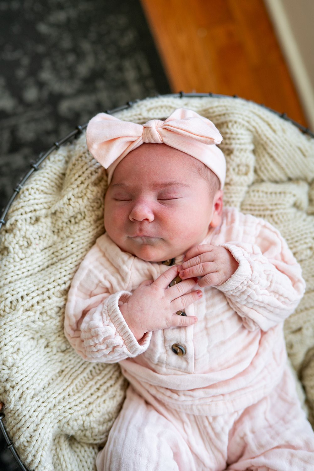 Sleeping newborn baby in pink outfit with matching bow rests peacefully on cream knit blanket.
