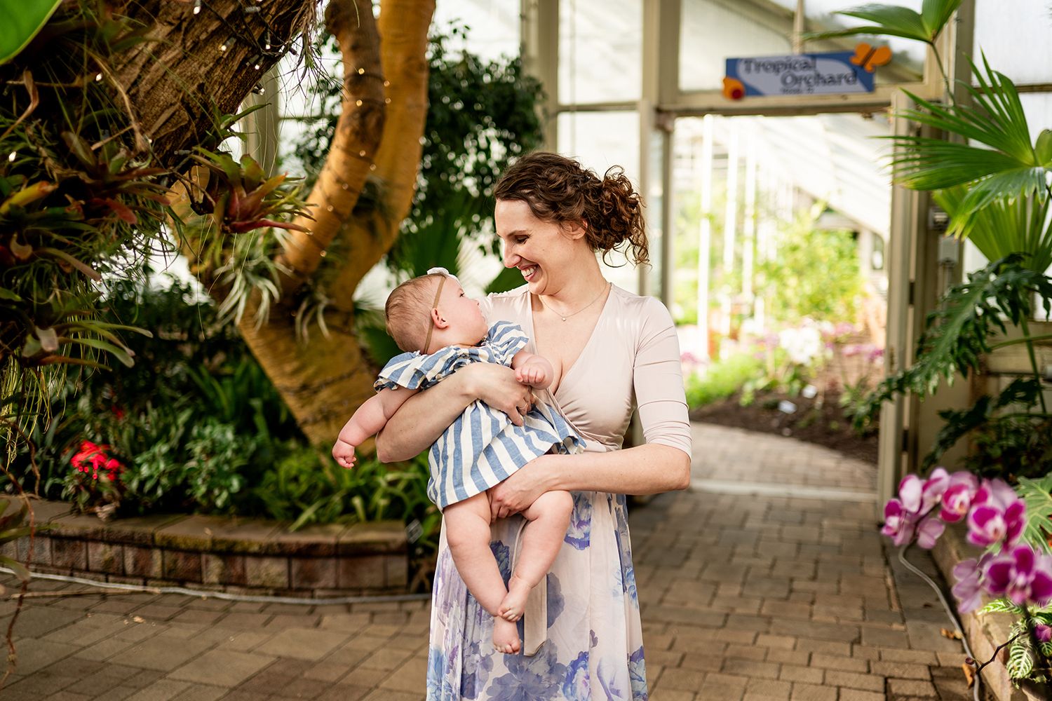 Two figures share a joyful moment together in a lush greenhouse setting with brick pathways and tropical plants.
