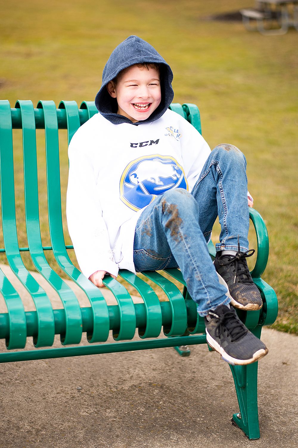 A joyful child wearing a white hoodie and blue cap relaxes on a green park bench on a sunny day.