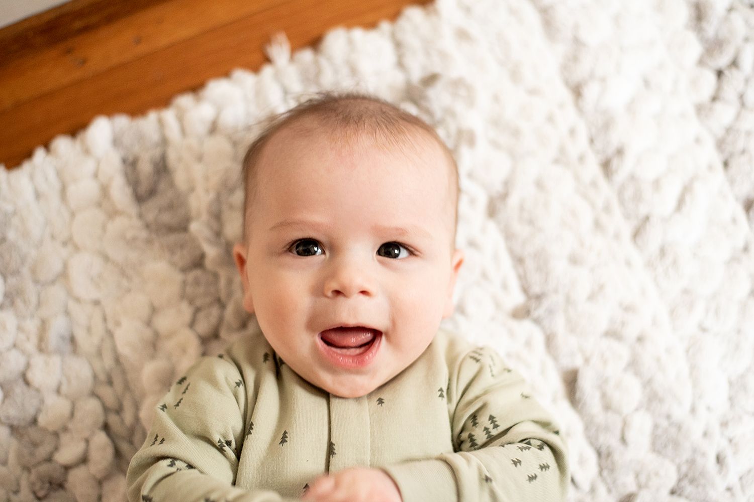 A happy baby in light green clothing lies on a fluffy white blanket smiling at the camera.