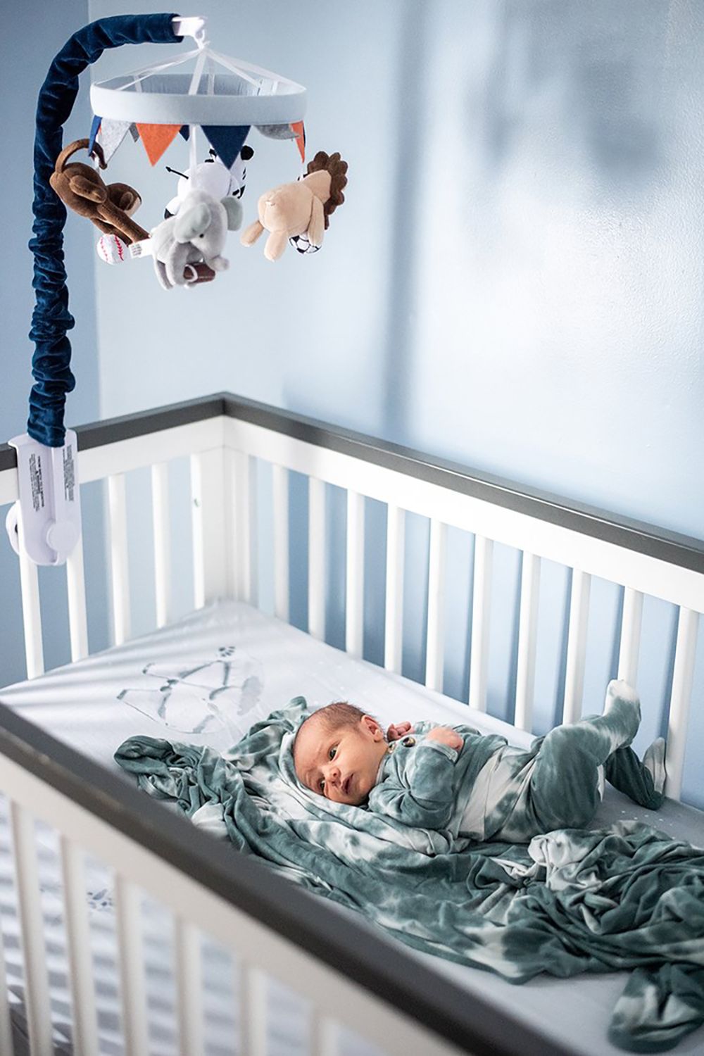 Baby in blue sleepwear lying in white crib with animal mobile hanging above.