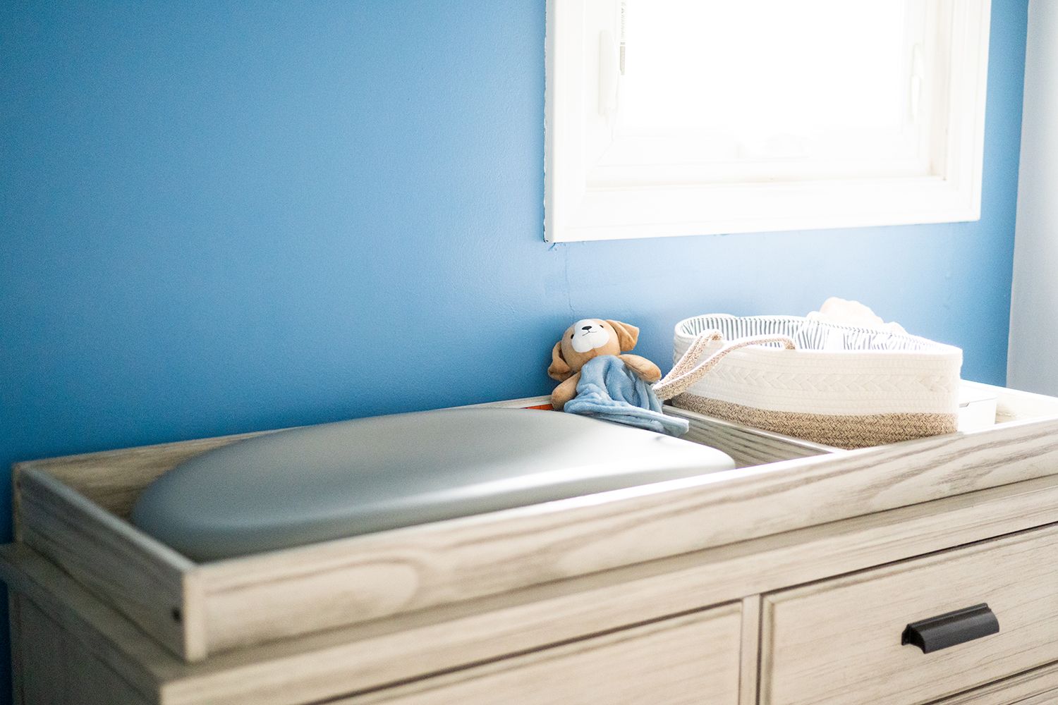 A white changing table dresser against a vivid blue wall with a plush toy and basket on top.