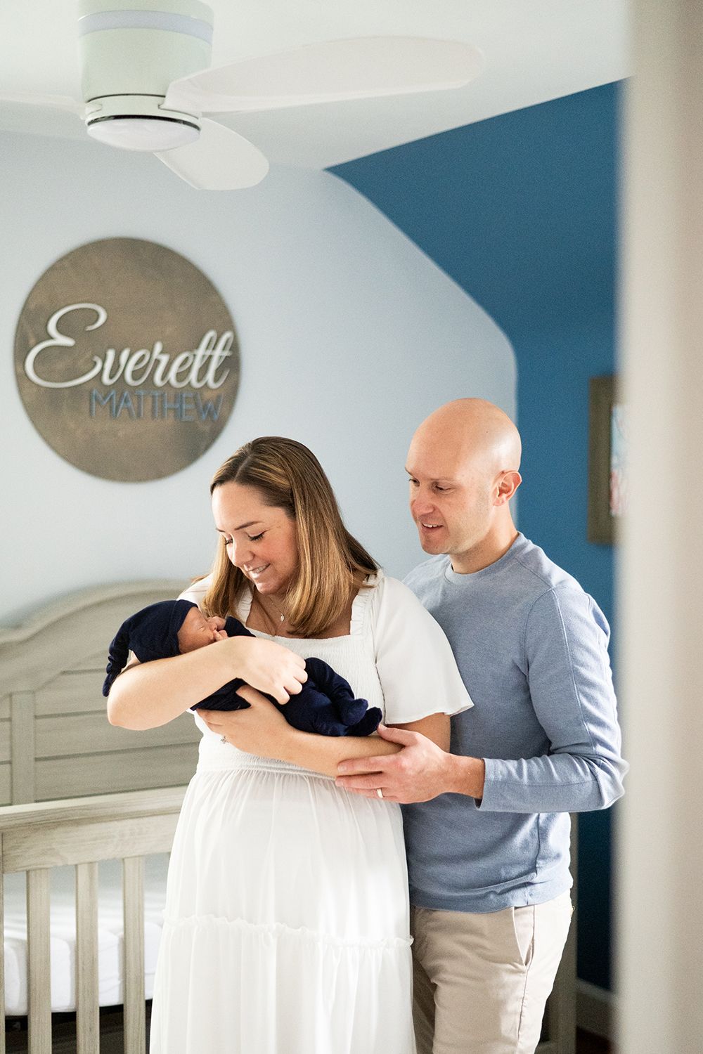 A couple stands in a nursery room with white furniture and blue accent wall while holding their newborn baby.