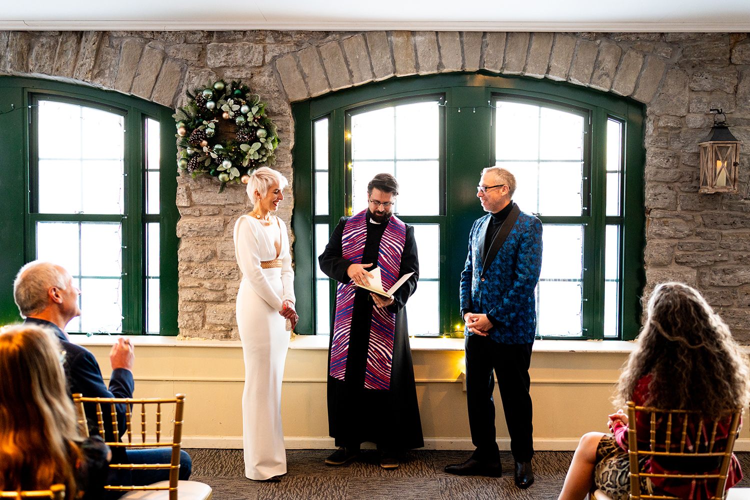 A small wedding ceremony takes place in front of arched windows with a holiday wreath in a stone wall venue.