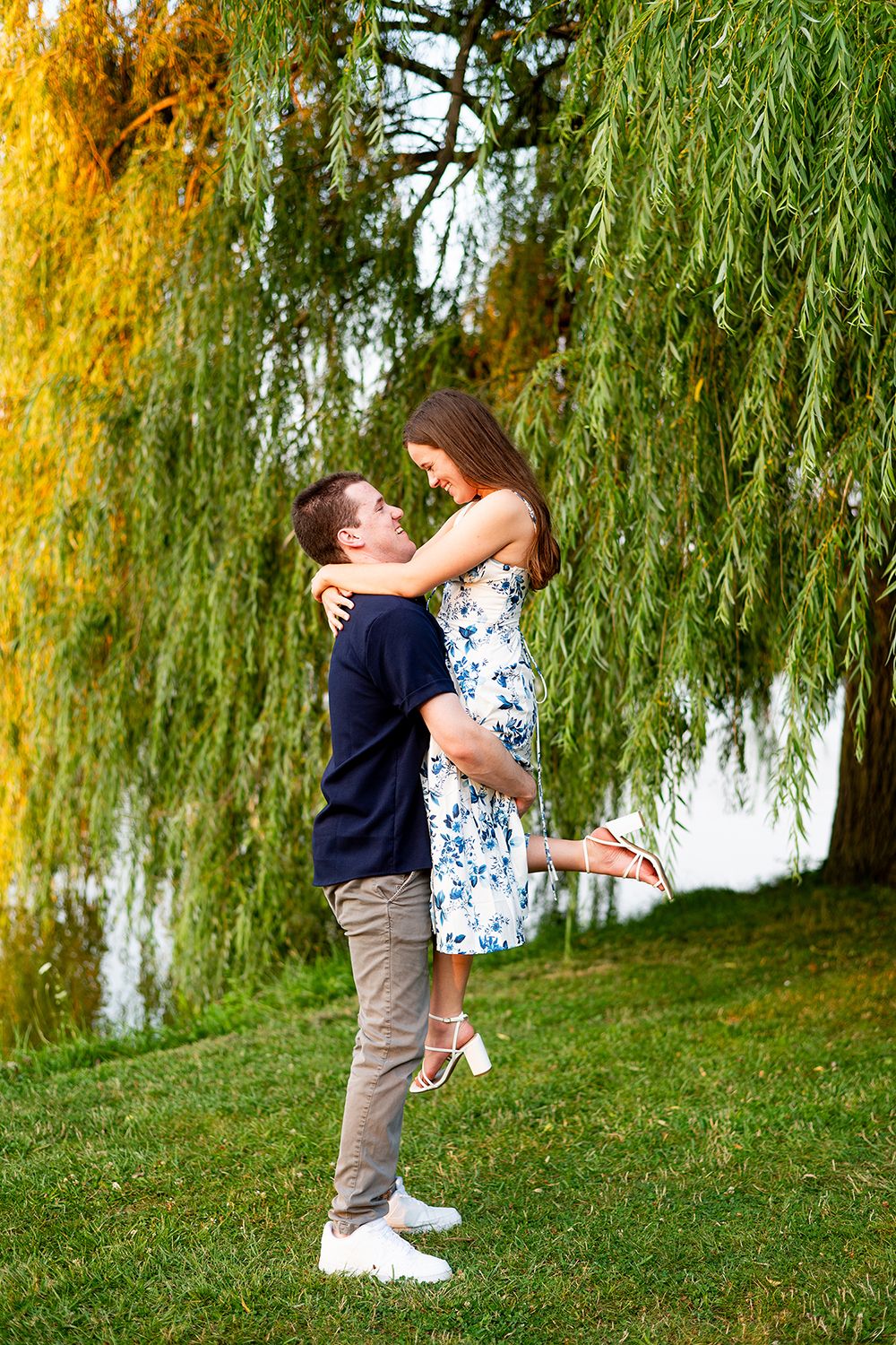 A romantic couple embraces on a grassy hill beneath cascading willow tree branches in autumn.
