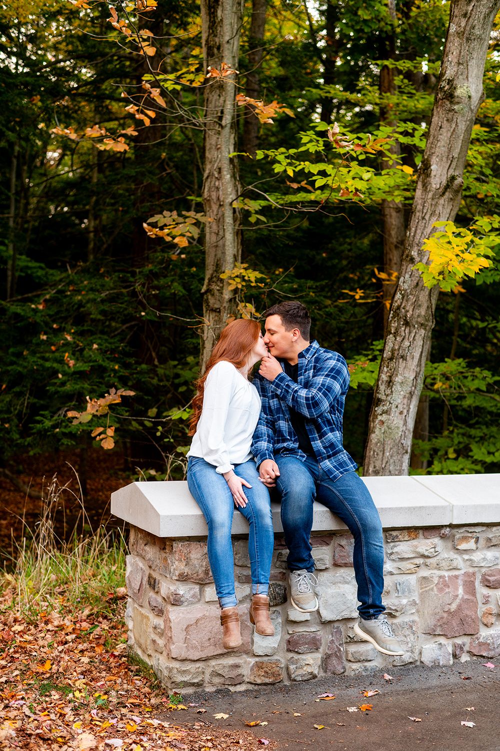 A couple sits together on a stone wall surrounded by autumn foliage and fallen leaves in a park setting.