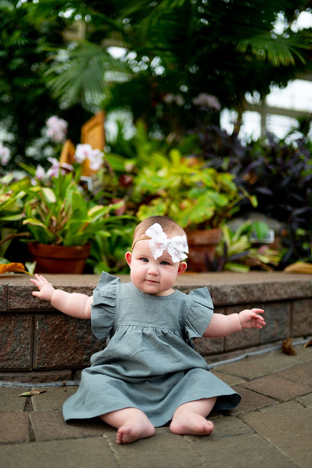 Baby wearing gray dress rests on brick patio surrounded by garden plants.