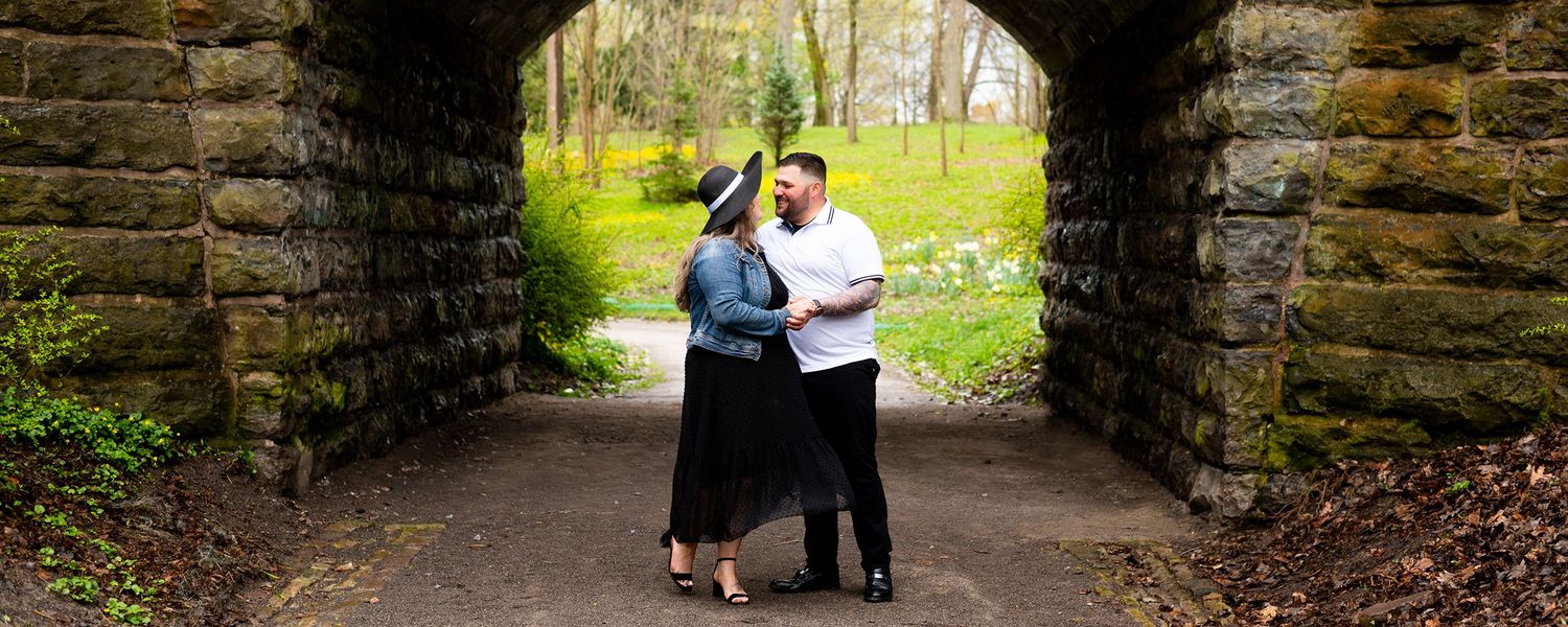 A romantic moment captured under a rustic stone archway in a park with greenery visible in the background.