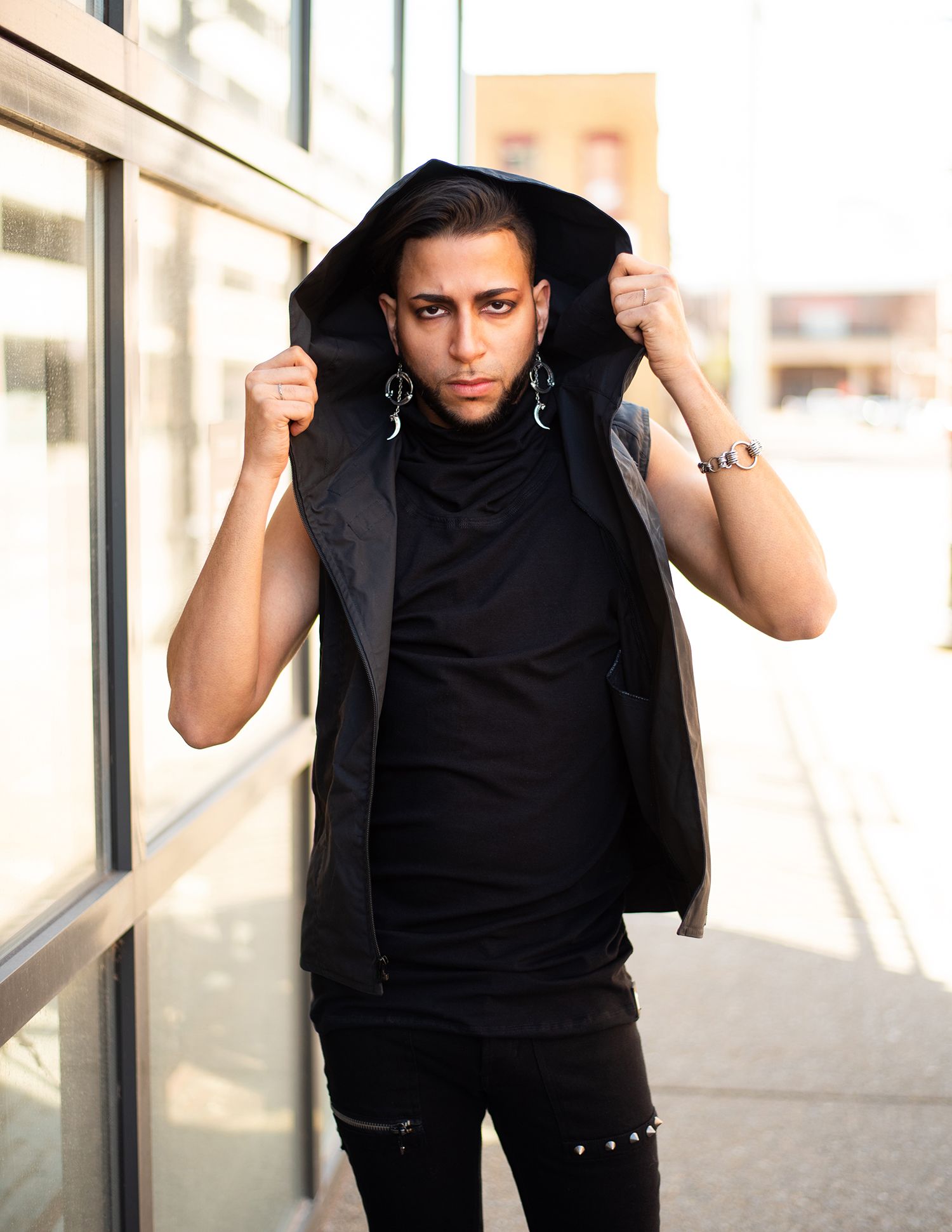 A model in monochromatic black attire poses against a wall while adjusting their hooded vest and wearing silver earrings.