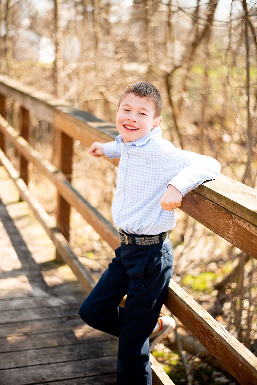 Child in blue gingham shirt and dark pants stands happily on wooden bridge walkway in autumn forest.