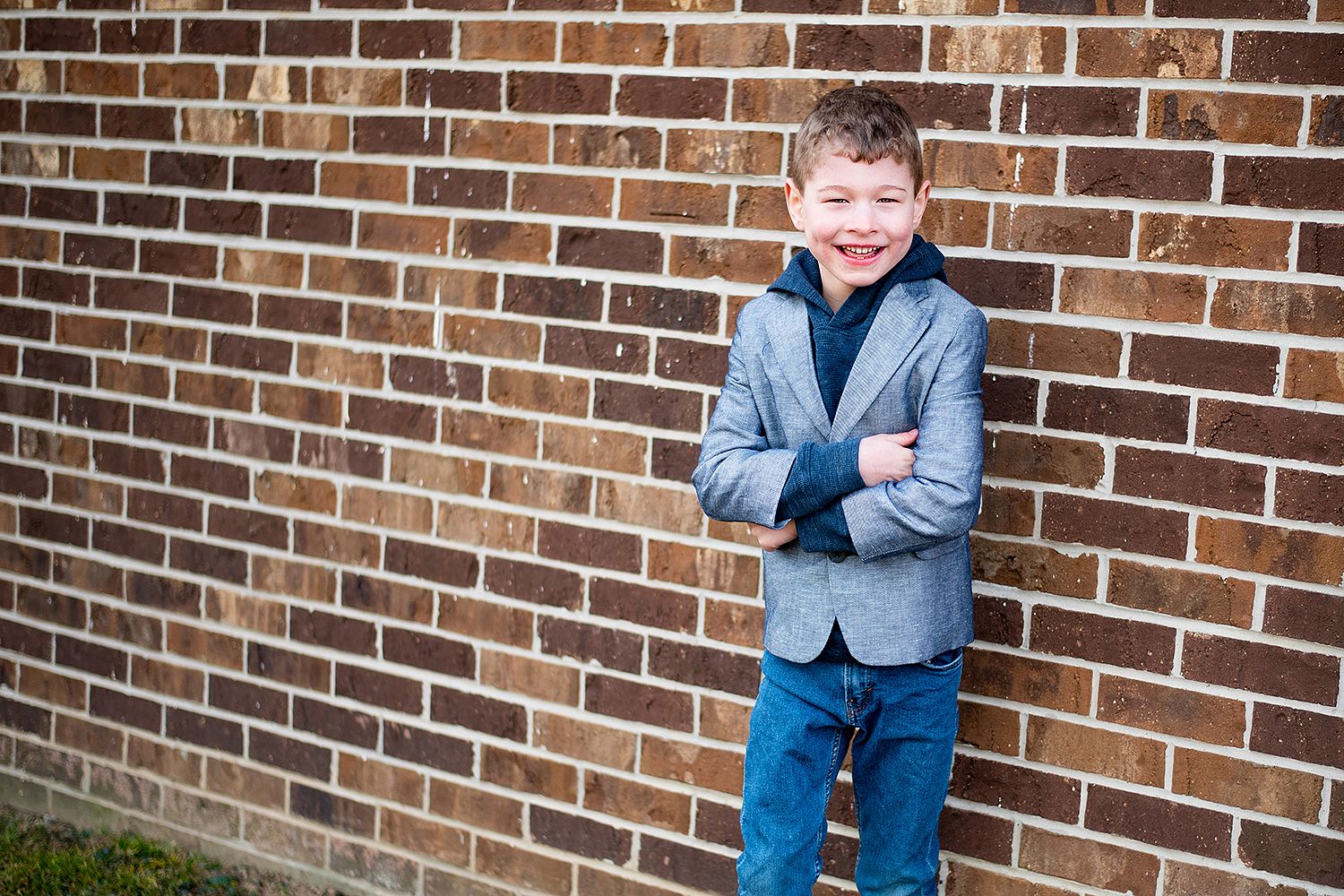 Young person in gray blazer and jeans stands confidently with crossed arms against brick wall outdoors.