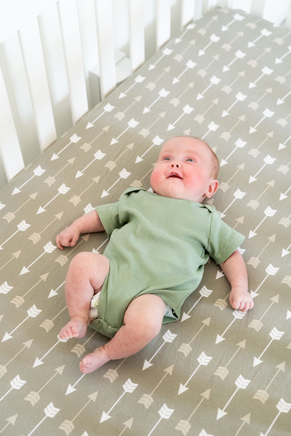A baby wearing a sage green onesie lies on a gray crib sheet with white arrow pattern.