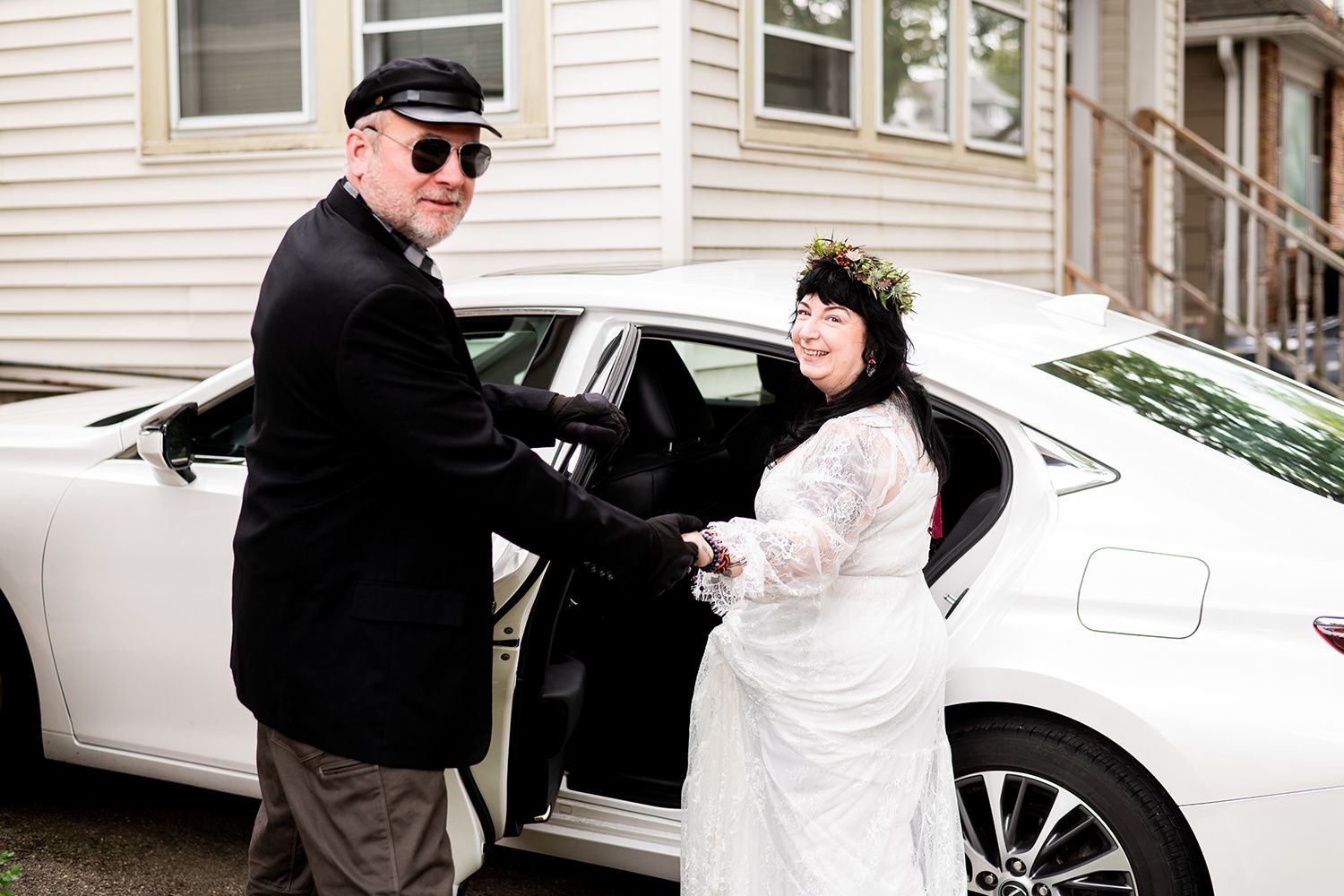 Two people in dark clothing interact beside a parked white car on a residential street.
