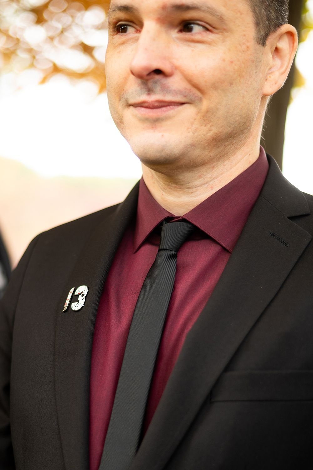 Professional headshot of someone wearing a black suit jacket with burgundy shirt and black tie.