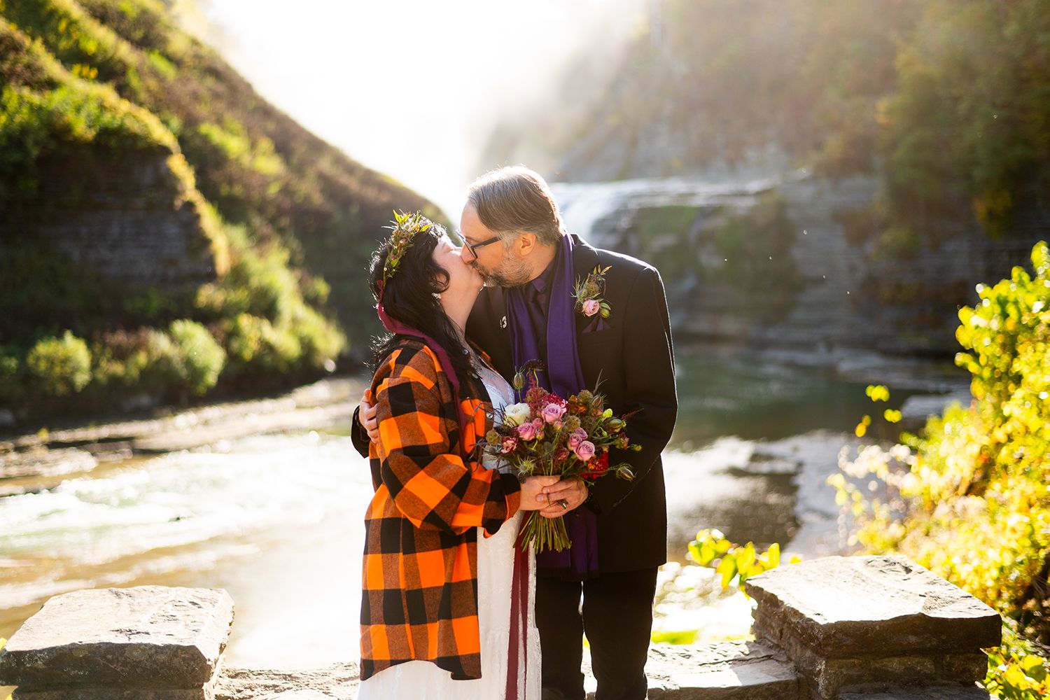 A couple wearing plaid and dark clothing embrace near a waterfall during golden hour, glowing in sunlit mist.