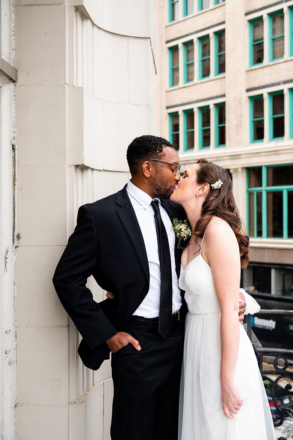 A romantic wedding couple share a tender kiss on a city street with turquoise windows in the background.
