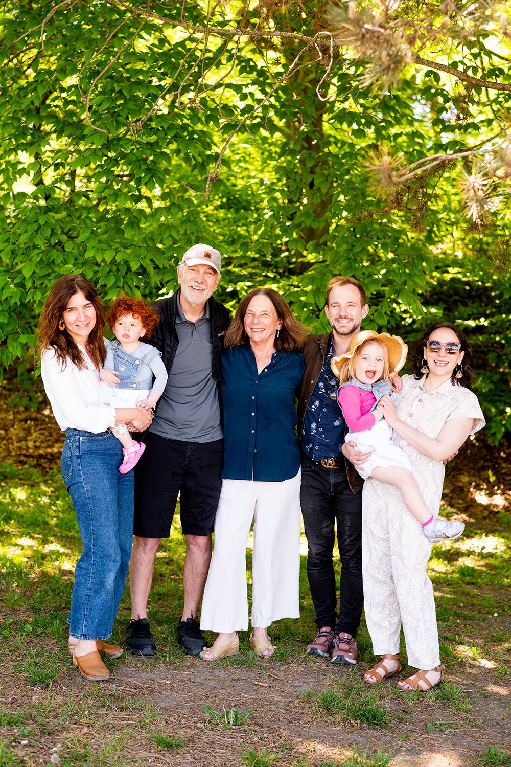 Extended family gathering for a group photo in a sunlit wooded area on a summer day.