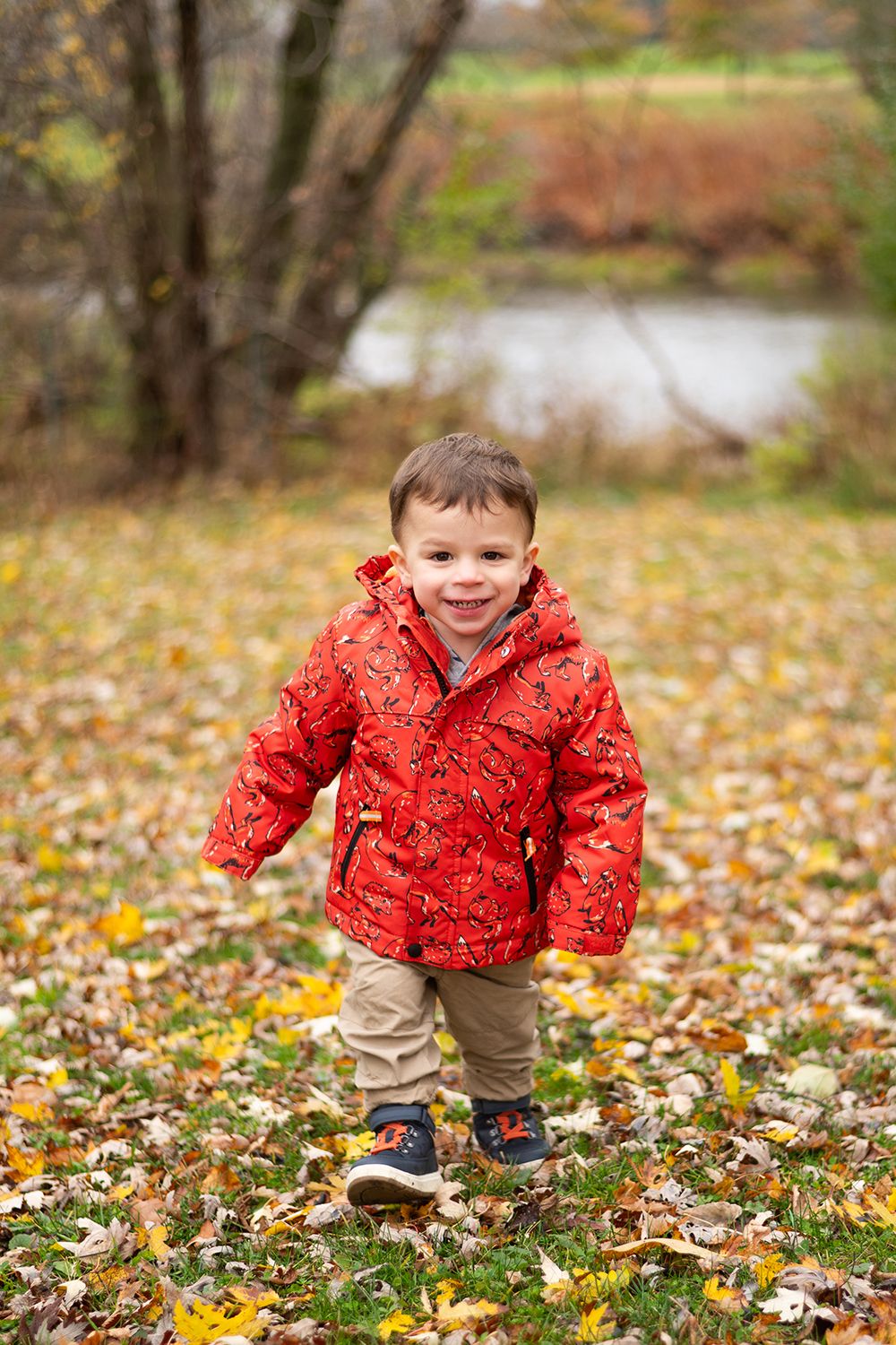 Young child in red patterned jacket plays in colorful autumn leaves by a pond on a fall day.