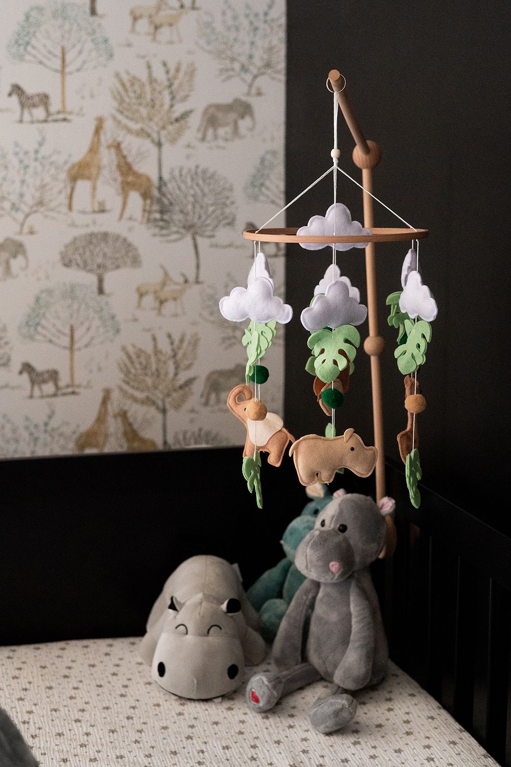 Safari-themed nursery mobile with plush animals and monstera leaves hangs above soft stuffed toys on a patterned surface.